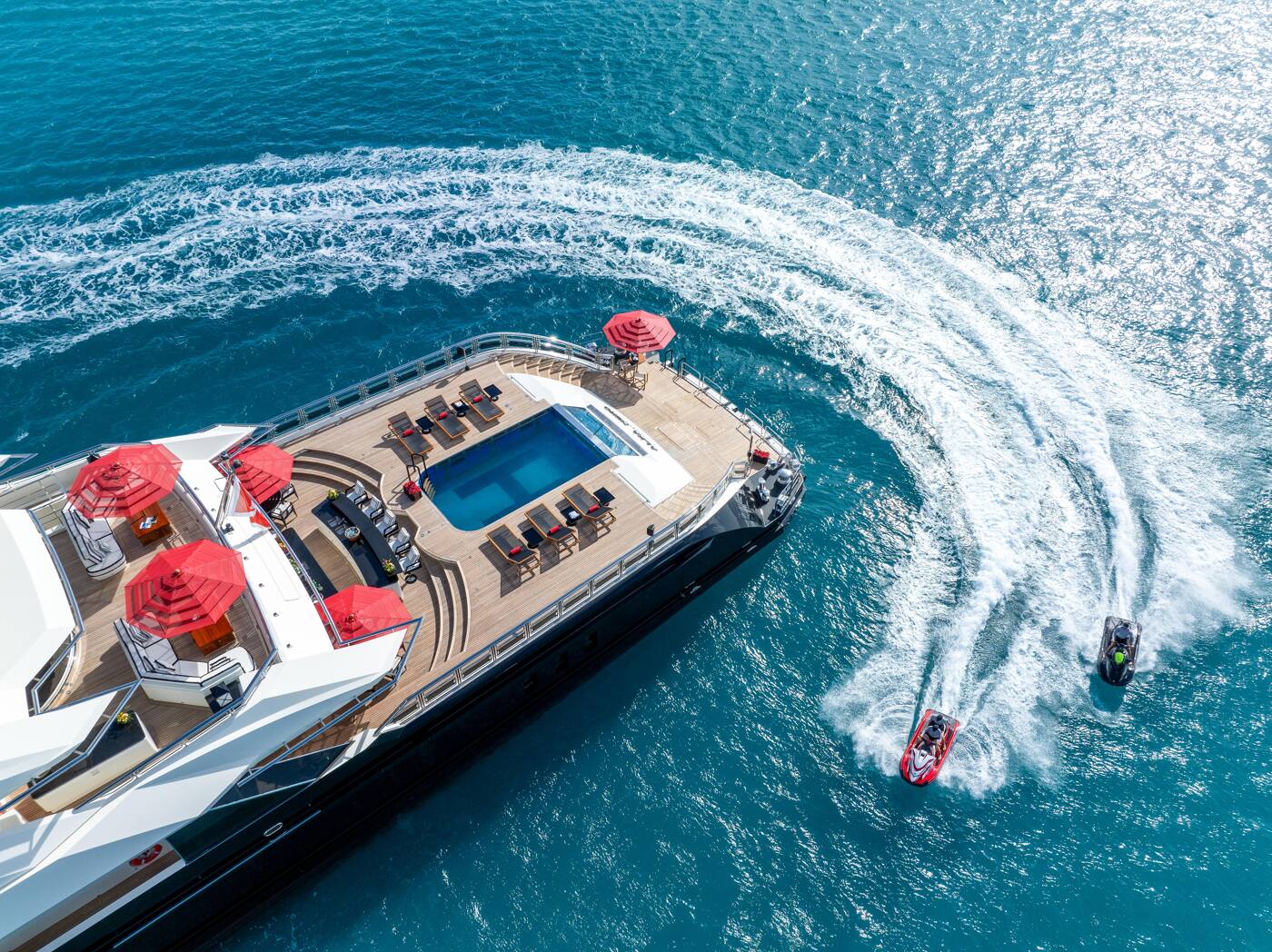 Aerial view of the aft deck pool and red parasols aboard ALFA NERO, two jet skis carving through her wake